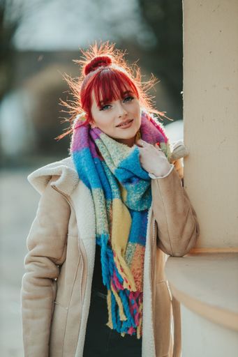 A portrait of a young red-haired girl with bangs holding her warm colorful scarf looking at the camera