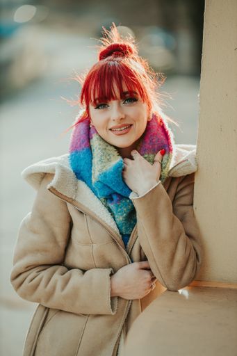 A portrait of a young red-haired girl with bangs holding her warm colorful scarf looking at the camera