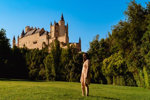 A young female posing against a historic Alcazar Castle surrounded by green trees in Segovia, Spain