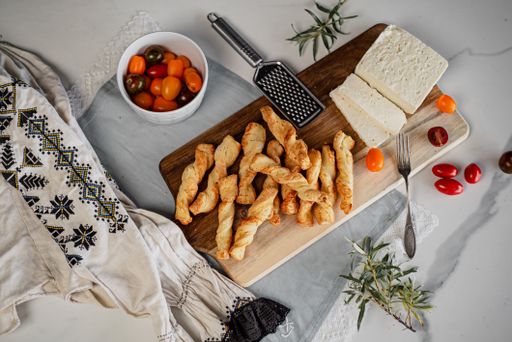 A pile of salted breadsticks on a wooden cutting board with some cheese and tomatoes