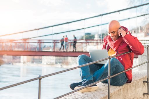 A bald Latin male talking on his phone and working with a laptop outdoors