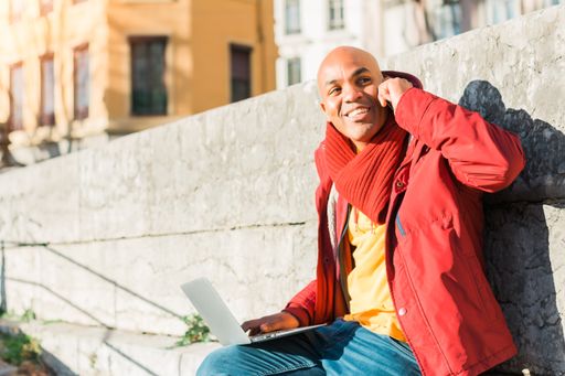 A bald Latin male talking on his phone and working with a laptop outdoors