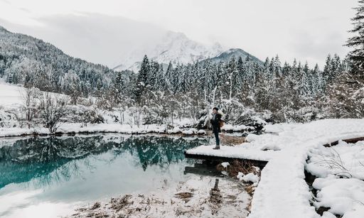 Young woman standing on path by green lake in winter. Deep snow, mountains, landscape. Lake Zelenci nature reserve, park. Kranjska gora, Slovenia.