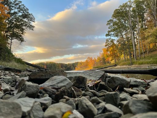 A tranquil autumnal scene featuring a mountain riverbank, with a setting sun in the background