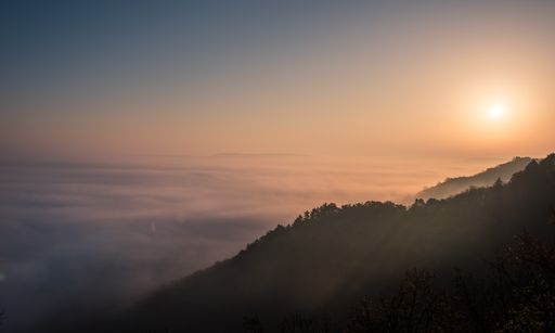 A beautiful shot of forested mountain above the clouds with the sun shining in the background in Hungary