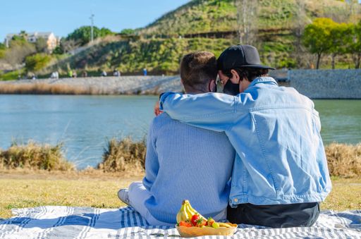 Picture of View from Behind of a Gay Couple Hugging Sitting in Tablecloth Picnic In front of a Lake in a Park