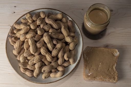 A top view of a bowl of peanuts in the shell, a glass jar, and a slice of bread with peanut butter