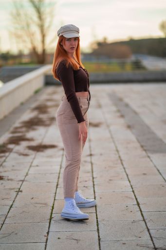 A vertical shot of a red-haired young woman in a hat posing in the park