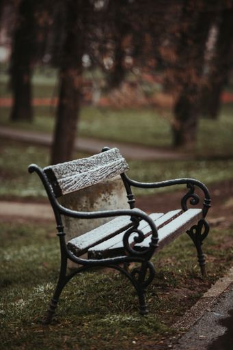 a vertical shot of a bench lightly covered with snow next to the trash on the blurred background