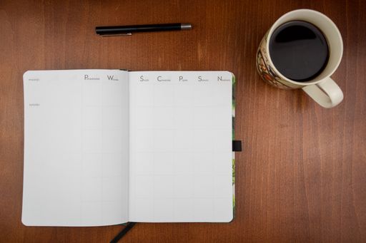 A top view of an open empty weekly dairy planner with a pen and coffee on an office wooden desk