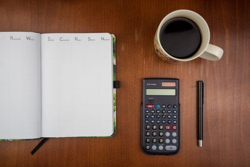 A top view of an open empty weekly dairy planner with office supplies and coffee on a wooden desk