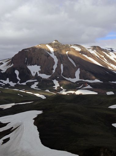 A vertical shot of a mountain covered in snowy patches under a cloudy sky