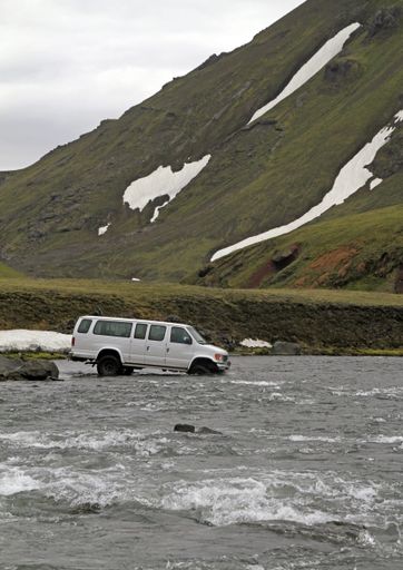 A vertical shot of an SUV car driving into the water near a mountain