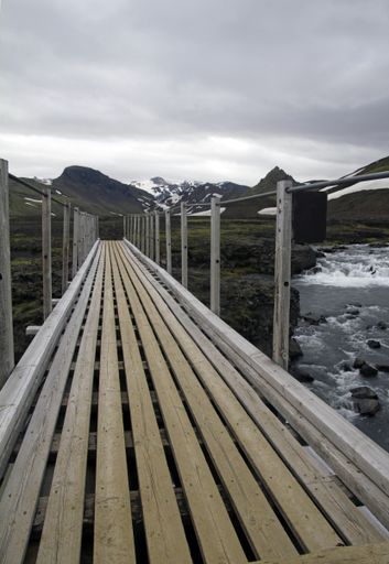 A vertical shot of a wooden bridge over a water stream under a cloudy sky