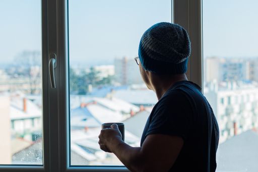 A young man drinking coffee and looking out the window at the snowy city