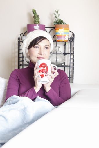 A vertical shot of a Hispanic woman in a hat sitting on a couch and drinking a hot coffee