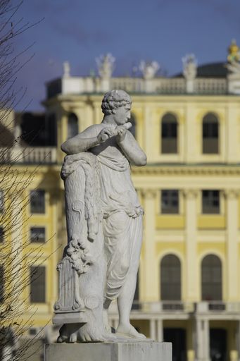 Figure is playing transverse flute. Stands in the main Park in front of Schoenbrunn Castle. The Castle glows yellow from behind.