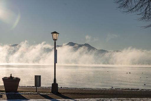 A street lamp on thpavement near a foggy lake on a sunny day
