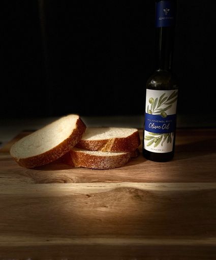 A photo of a glass bottle of healthy Polyphenol Olive Oil lit up with sliced loaf of bread on cutting board with dark background