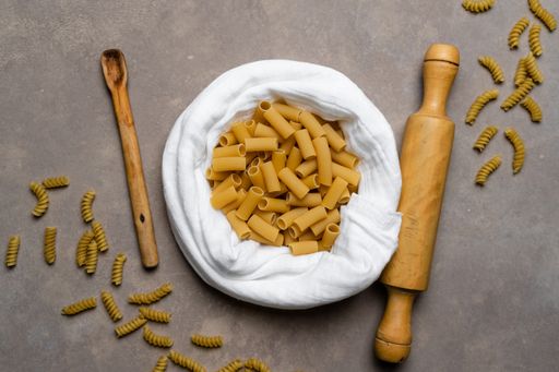 A top view of dry rigatoni pasta with a wooden spoon, rolling pin and white cloth