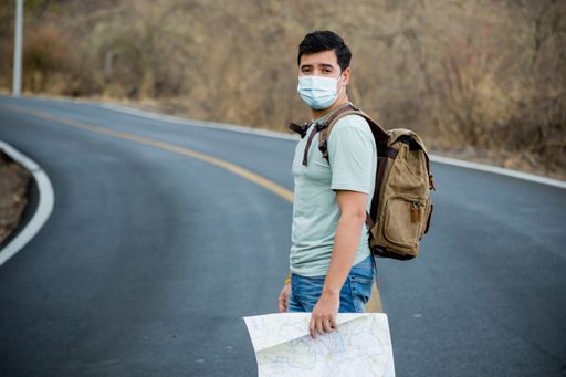A male traveler with a backpack and maps wearing a sanitary mask standing on the road