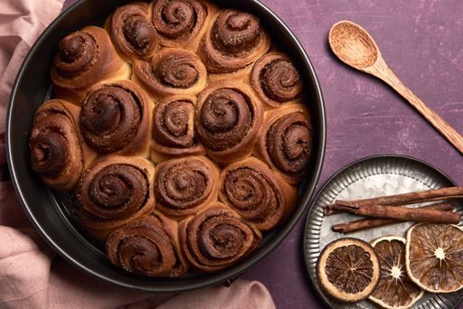 A high angle shot of fresh cinnamon roll buns with a wooden spoon, cinnamon, and dried oranges