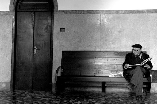 A grayscale shot of an elderly man sitting on a bench reading a book indoors in Parma, Italy