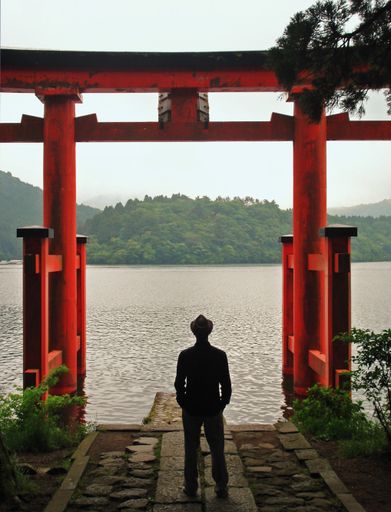 A vertical shot of a male in black standing in front of the Lake Ashi in Hakone, Japan