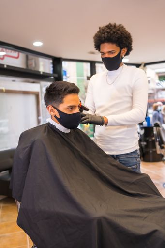 A male hairdresser with a mask cutting the hair of a young man