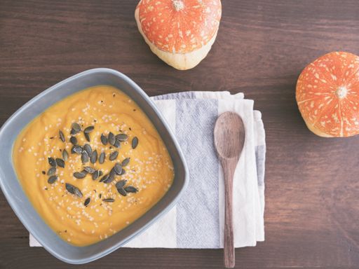 A bowl of pumpkin soup with two pumpkins and a wooden spoon on a wooden table