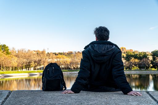 Traveler man in a black coat and backpack, sitting in front of a pond while resting.