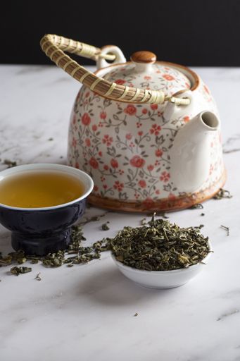 A vertical selective focus shot of green leaf tea, a container, and kettle on marble background