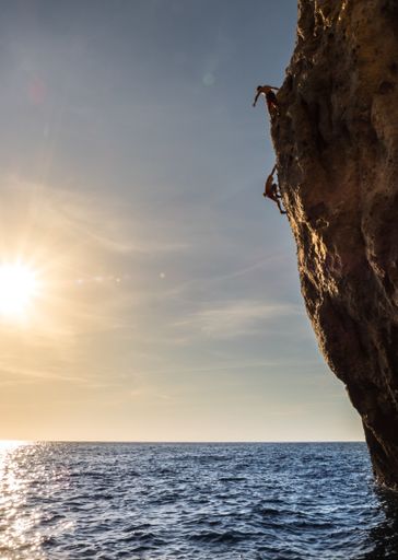 A vertical shot of two guys climbing down a cliff over the blue sea on a sunny day