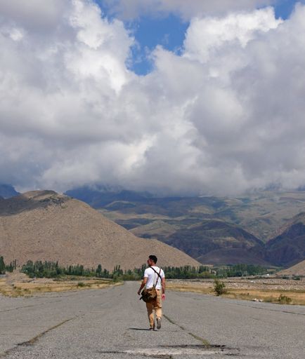 A vertical shot of a man walking on the road around the mountains under a cloudy sky