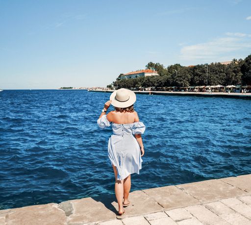 A shallow focus of a female wearing a light blue dress and a floppy hat and posing near the sea