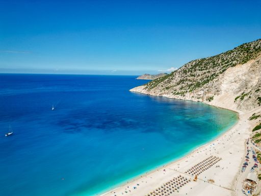 Drone shot of Myrtos beach hillside, Kefalonia, Greece with parking, umbrellas and a yacht in the cove