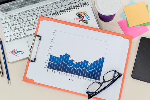 A top view of a laptop with financial data charts, glasses, paper clips, and a notebook on it in the office