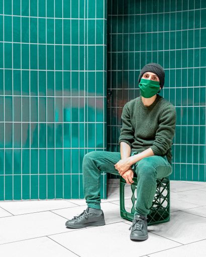 A man wearing a green face mask seated in front of a green brick wall