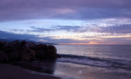 A picturesque scene of the purple toned sunset above sea waters seen through the rocky shore