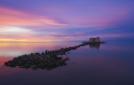 A wide angle shot of a small island in the middle of the ocean under a sky painted with multiple colors