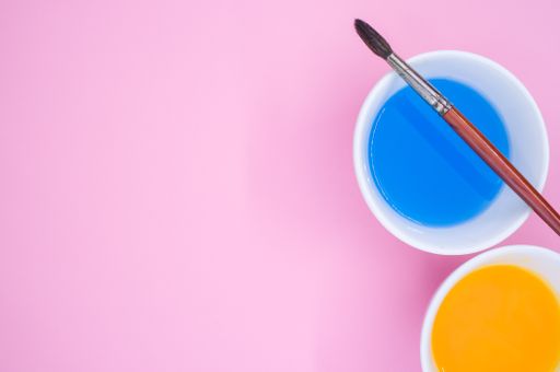 A top view of blue and yellow liquid watercolor paints on a bowl and paintbrush isolated on pink background