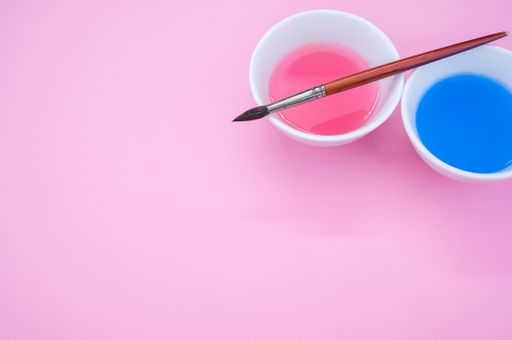 A high angle shot of pink and blue liquid watercolor paint on a bowl and paintbrush isolated on pink background
