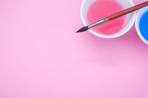 A high angle shot of pink and blue liquid watercolor paint on a bowl and paintbrush isolated on pink background