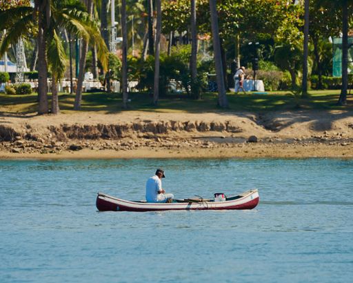 A closeup of a  man in a small boat sailing on a tranquil lake surrounded and a beach in the background