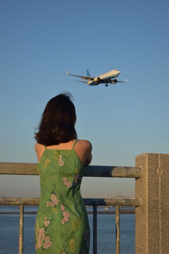 A woman with green dress standing under the flying plane in the blue sky.