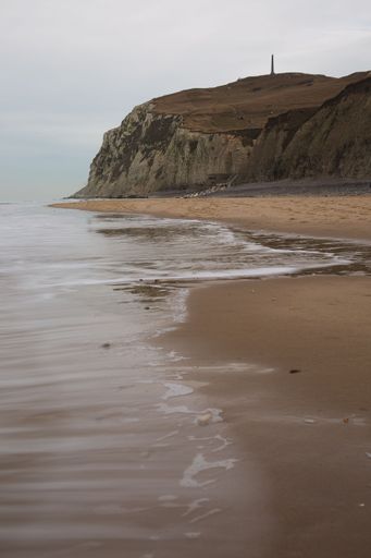 A vertical shot of the sea with long exposure surrounded by cliffs under a cloudy sky