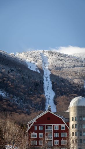 A wooden mountain lodge in the snowy terrain of Vermont surrounded by a ski mountain and a ski lift, with fresh snow covering the ground
