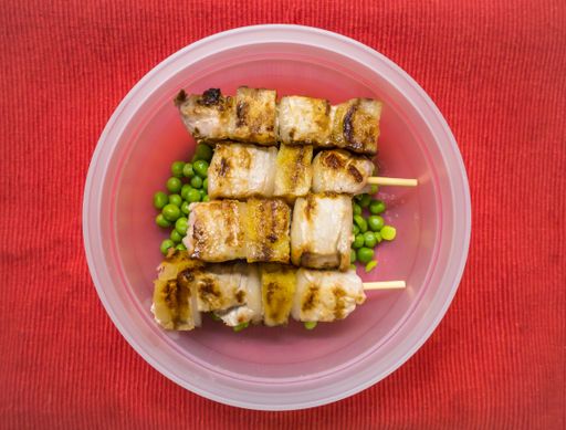 An overhead shot of pork on skewers with green beans in a plastic bowl on a red surface