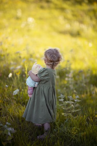 A vertical shot of a little child holding a doll while standing on a grass-covered field