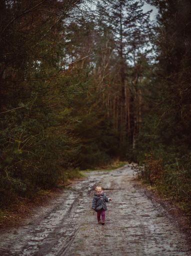 A vertical shot of a little child walking on a muddy road surrounded by trees